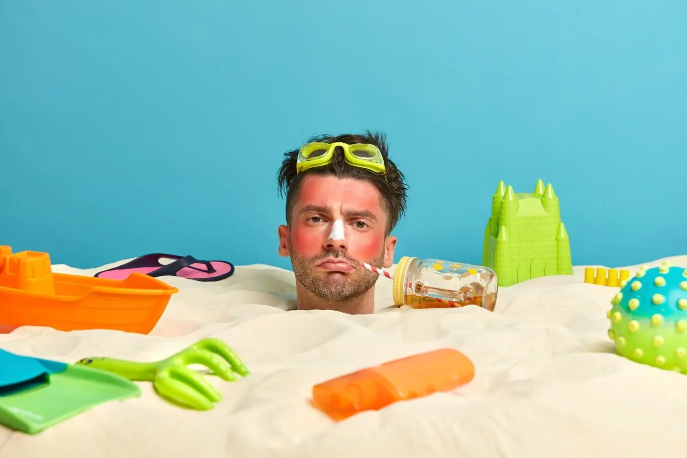 Kopf eines jungen Mannes mit Sonnencreme im Gesicht, umgeben von Strandaccessoires.